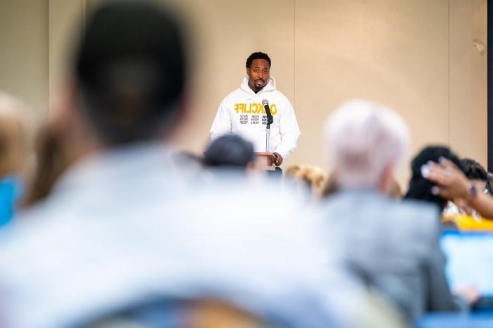 A speaker wearing a white "Oak Cliff" hoodie stands at a podium, addressing an audience.