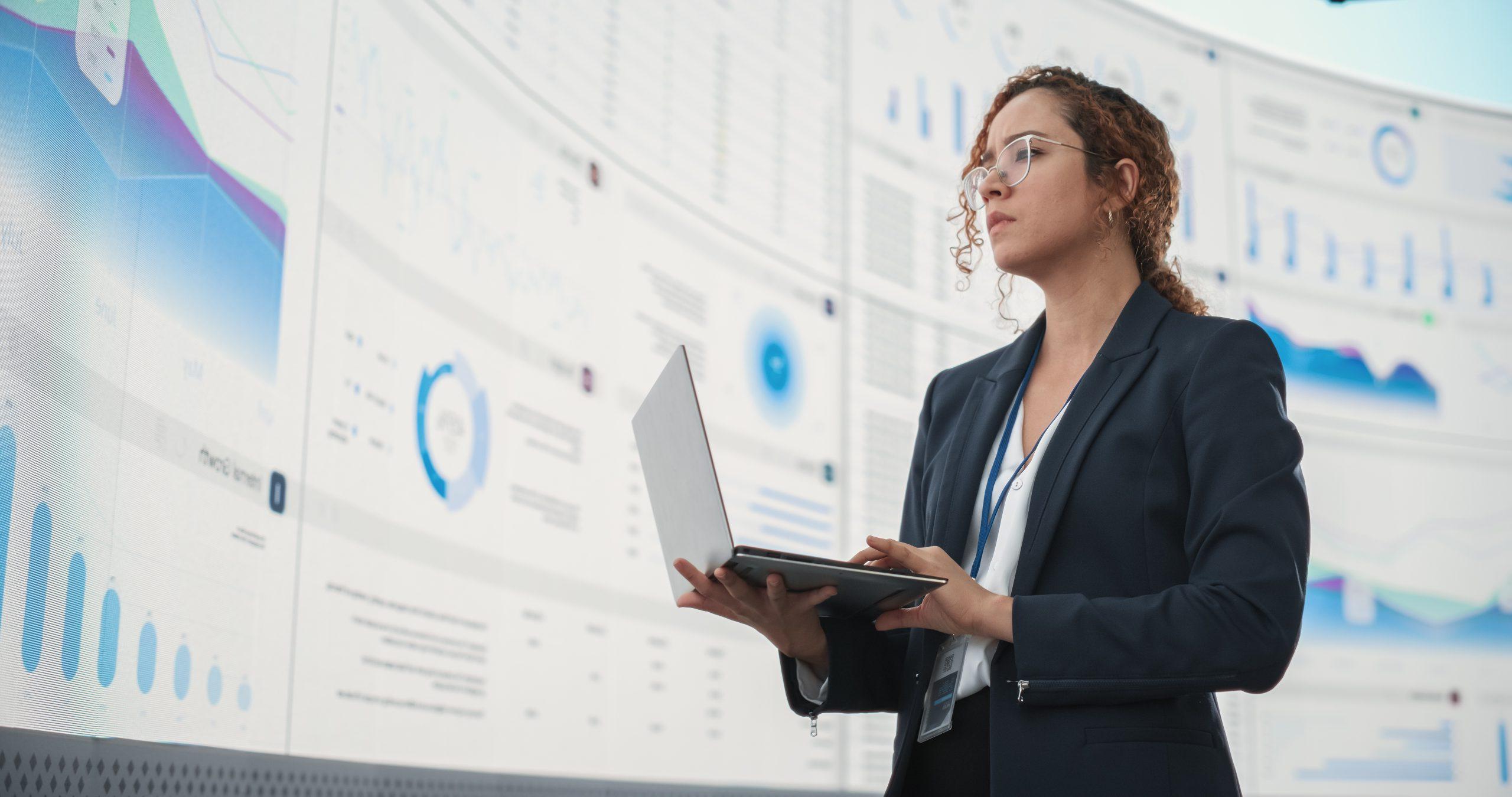 Successful Hispanic Businesswoman Holding Laptop Computer And Reviewing Financial Reports On Digital Screen In Monitoring Office. Female Leader Running Deep Learning, Artificial Intelligence Company.