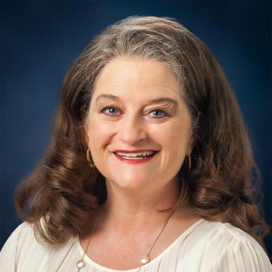 Portrait of a smiling woman with long curly hair, wearing a white blouse and gold earrings. Professional headshot with a dark blue background.