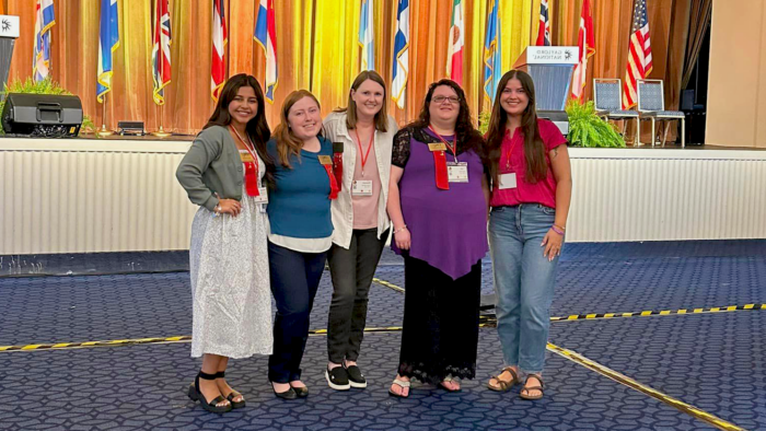 Four smiling female college students pose with a female faculty member in the middle. A stage behind them is adorned with flags representing countries around the world.